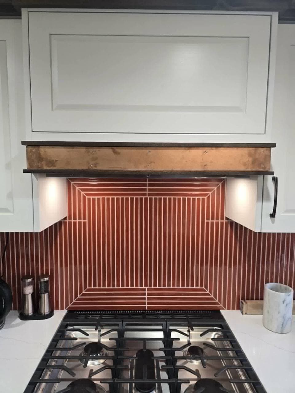 Raw bronze band wrapping the base of a range hood above a dark red vertical-stripe tile backsplash.