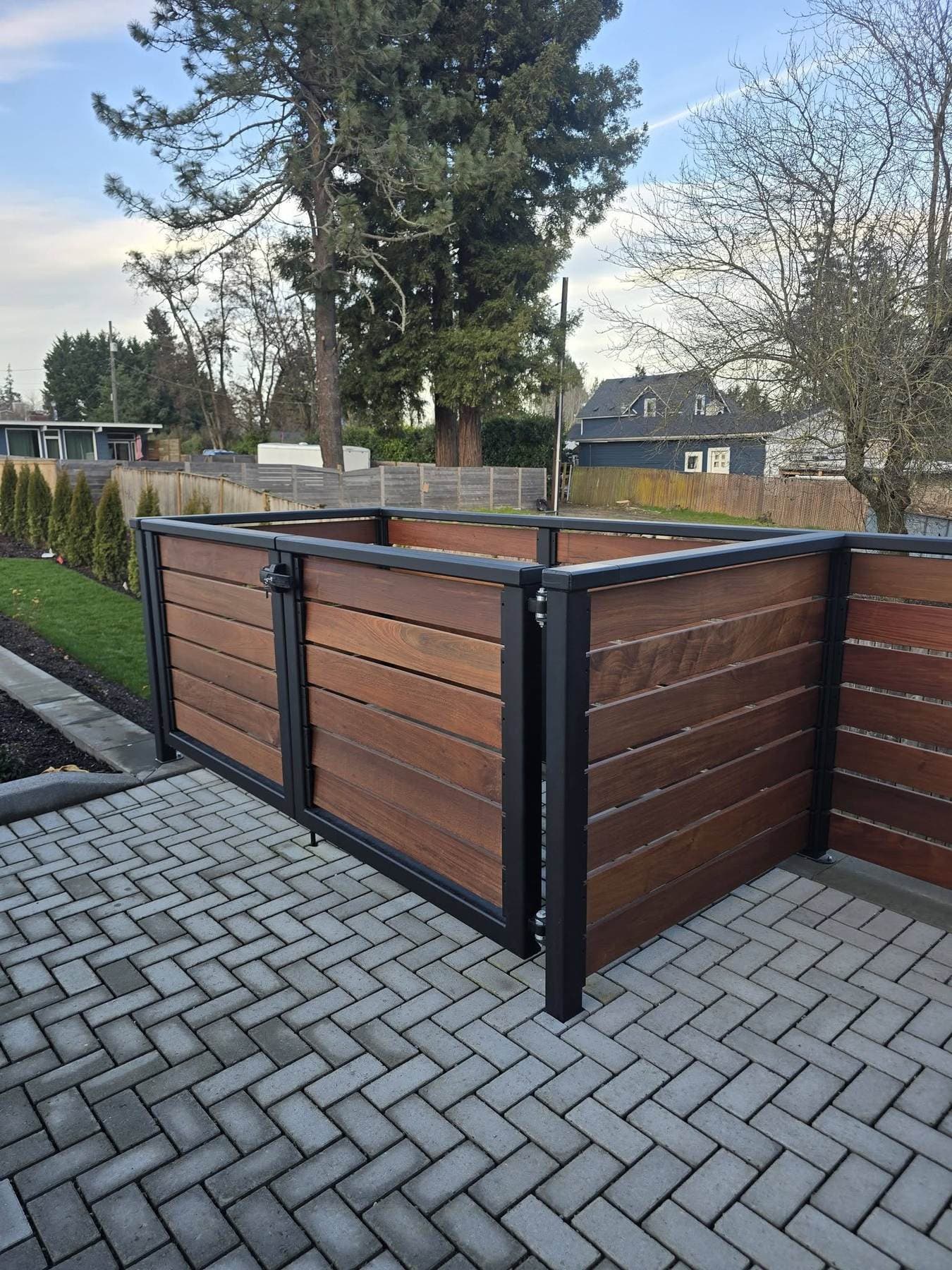 Steel-framed enclosure with horizontal wood slats and a locking double gate, on a herringbone paver patio.