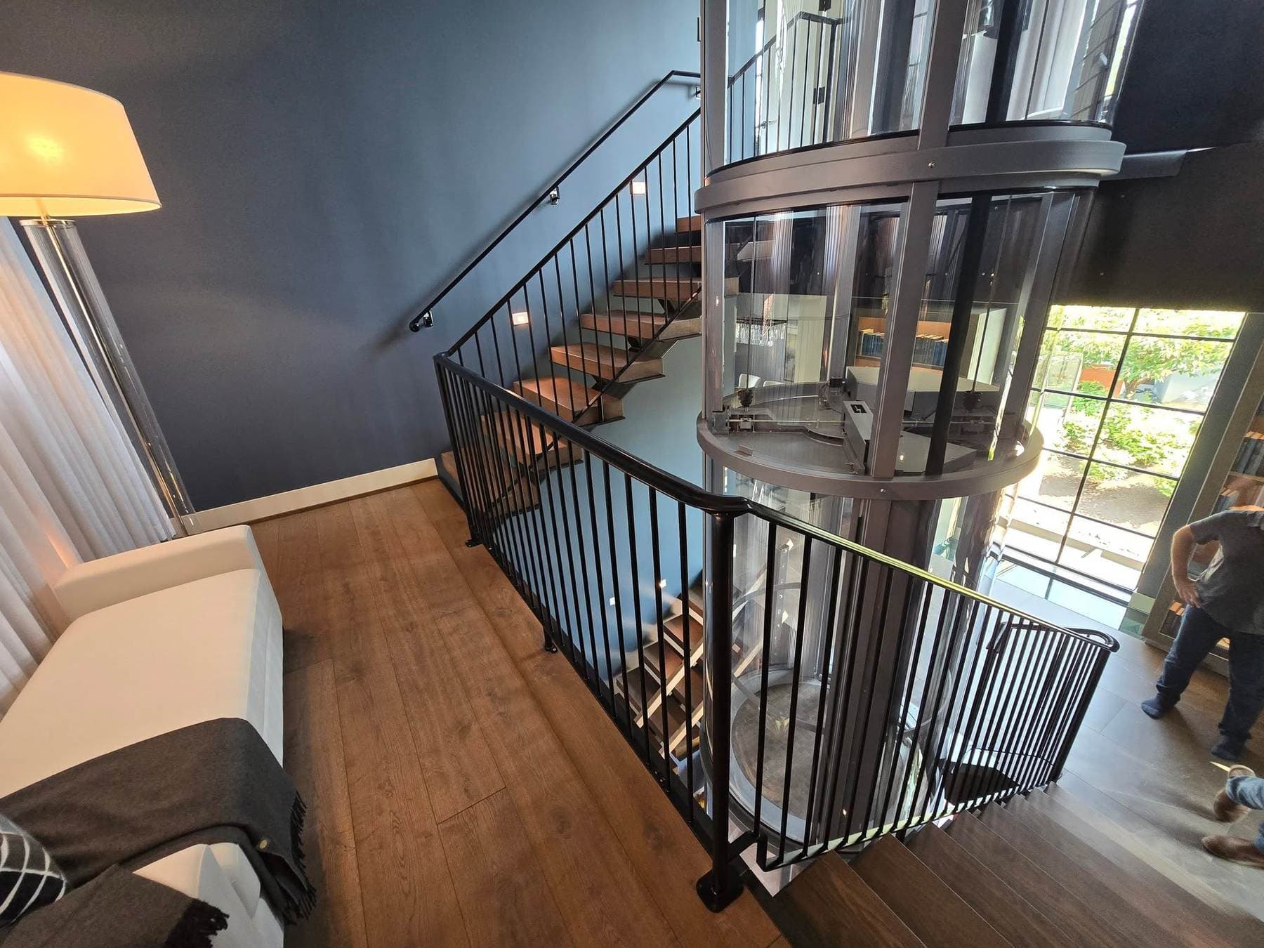 Black vertical-baluster stair railing wrapping a landing beside a glass elevator shaft in a multi-story home.