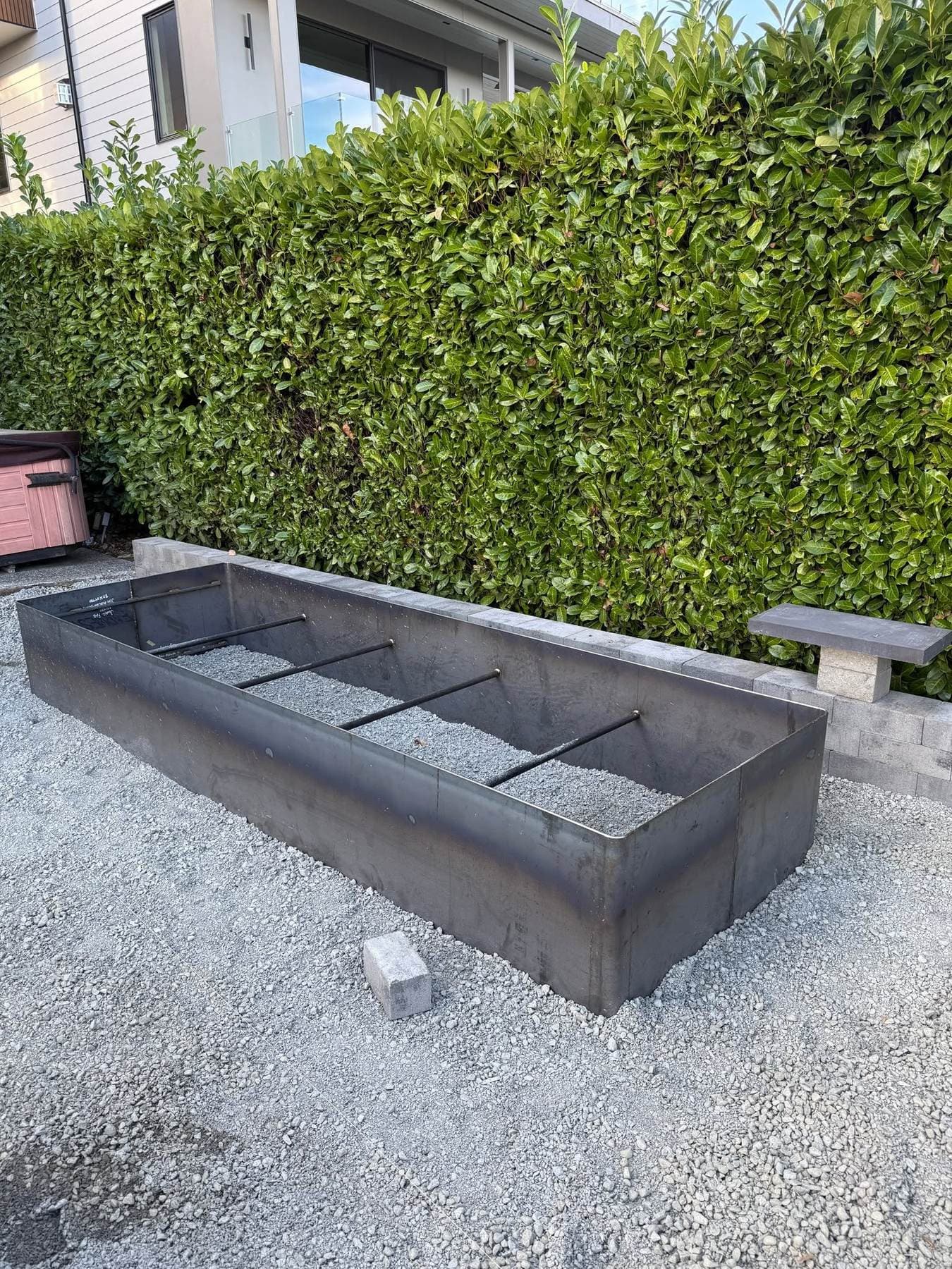Finished raw-steel planter box set against a hedge, with a paver border and stone bench nearby.