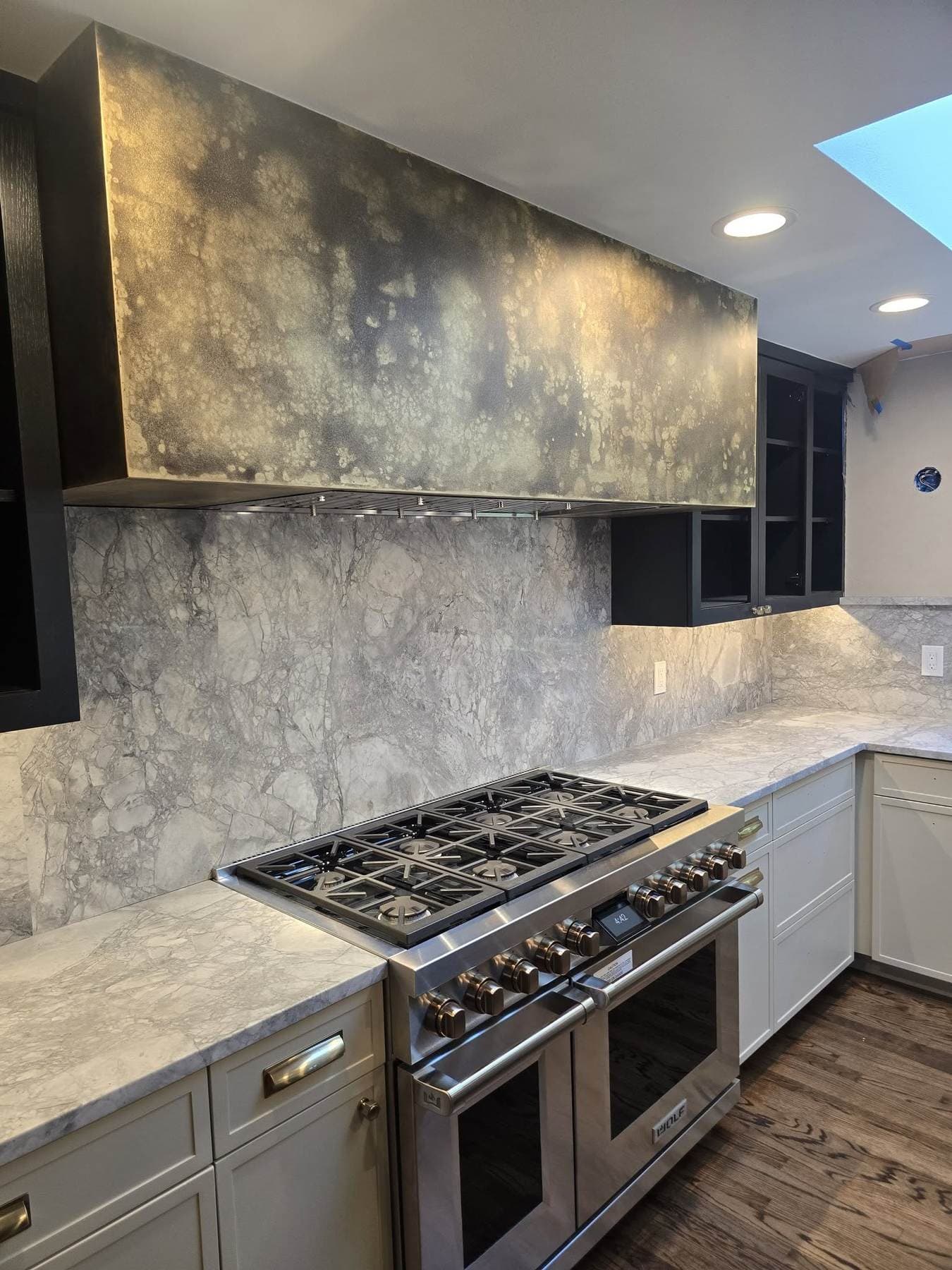 Large custom steel range hood with a mottled black-and-gold patinated finish above a professional range, set against a marble slab backsplash.