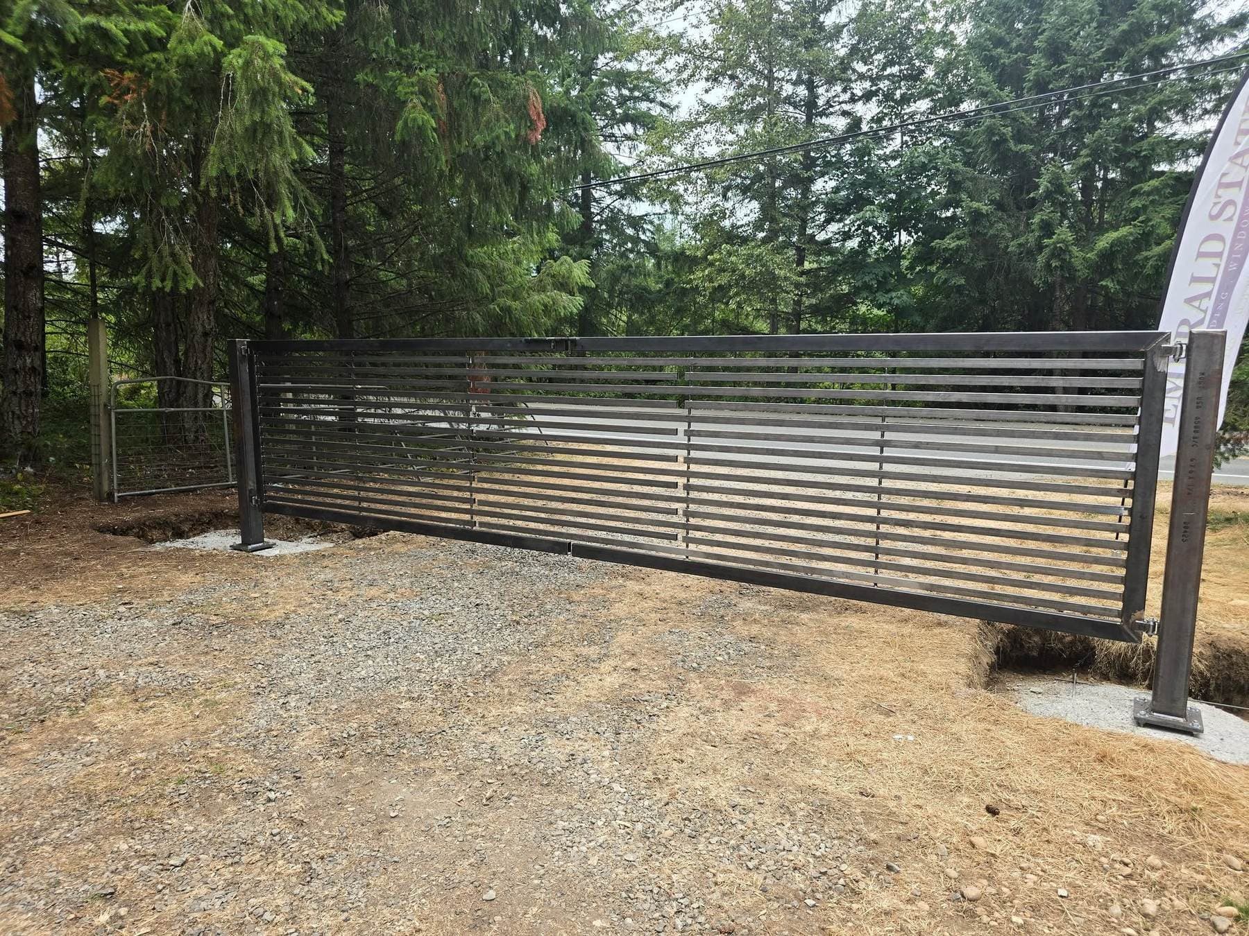 Wide sliding steel driveway gate with horizontal slats, set between concrete posts on a wooded rural property.