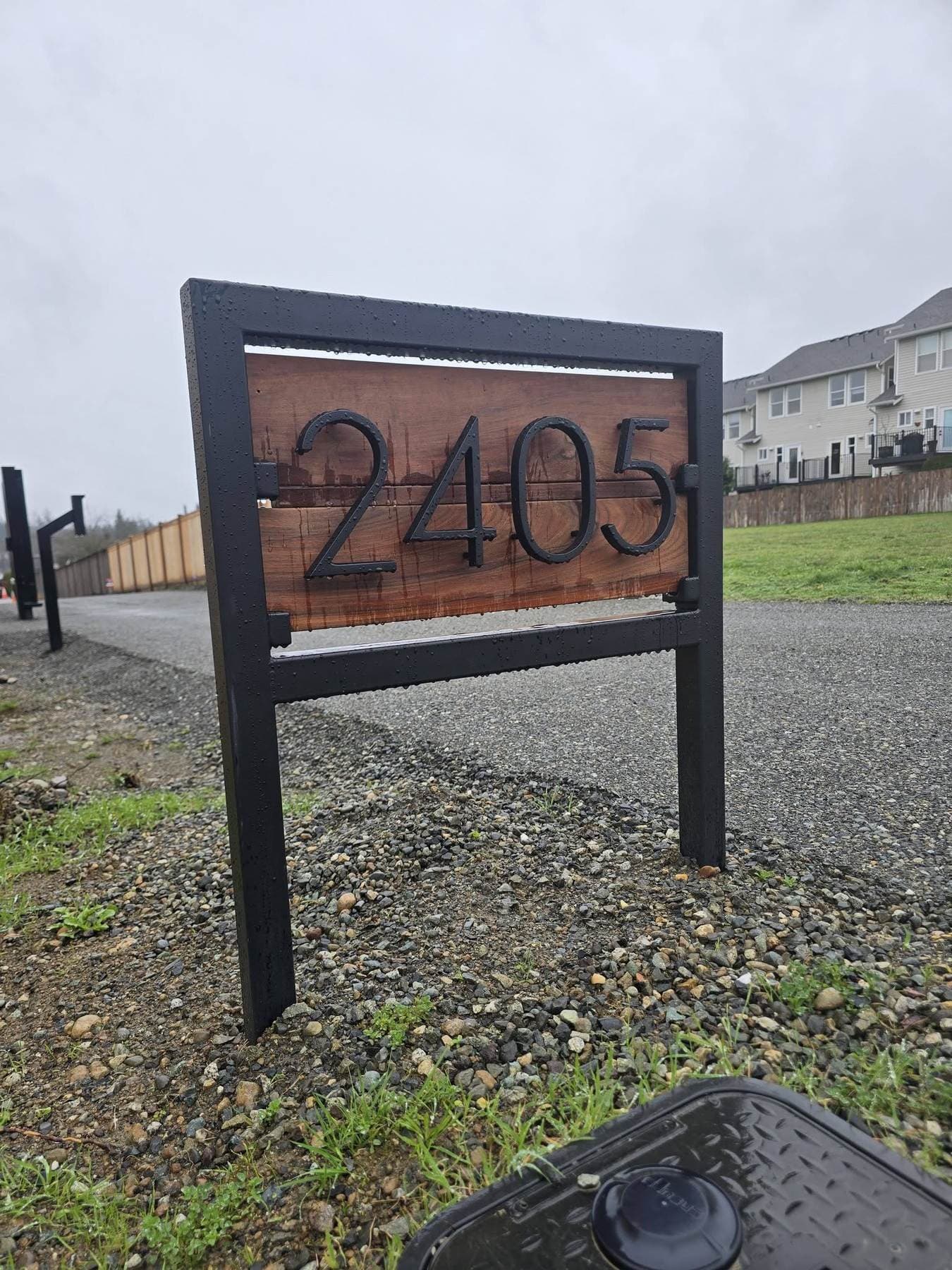 Framed steel-and-wood address sign with applied metal house numbers, mounted on a freestanding post.